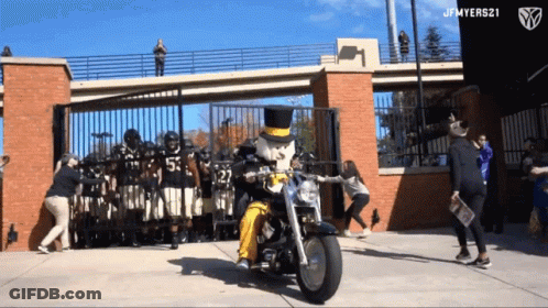 Man In Top Hat Riding Motorcycle Past Football Players GIF
