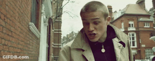 Man In Trench Coat Walking Down Street Next To Brick Building GIF