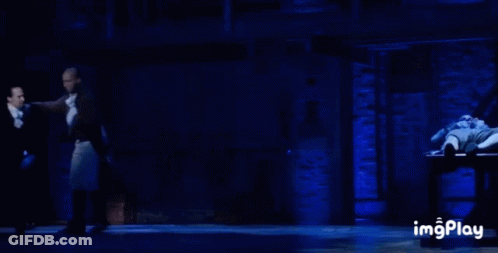 Man In Tuxedo Dancing On Stage In Front Of Man Laying On Table GIF