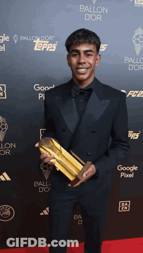 Man In Tuxedo Holding Gold Trophy In Front Of Ballon D'Or Wall GIF