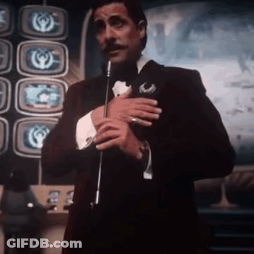 Man In Tuxedo Holding Microphone In Front Of Televisions GIF