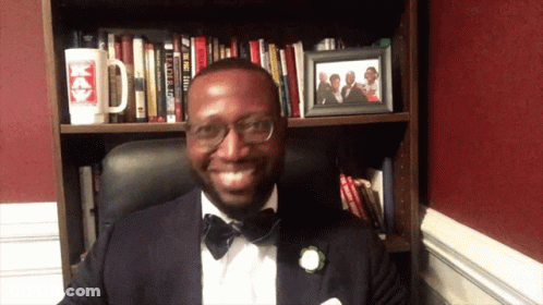 Man In Tuxedo In Front Of Bookshelf With Picture Of Man In Tuxedo GIF