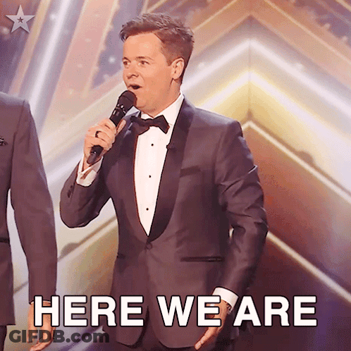 Man In Tuxedo Singing Into Microphone Here We Are GIF