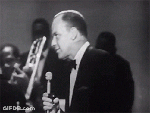 Man In Tuxedo Singing Into Microphone With Band Behind GIF