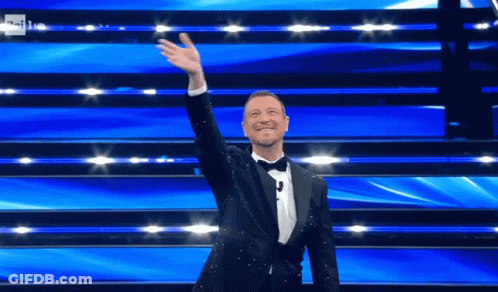 Man In Tuxedo Standing On Stage With Hand Up GIF