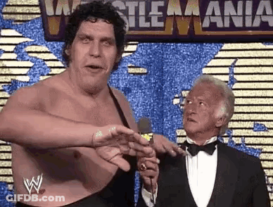 Man In Tuxedo Talking Into Microphone Beside Large Wrestler GIF