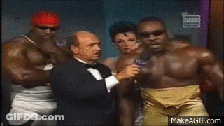 Man In Tuxedo Talking Into Microphone Beside Wrestlers GIF