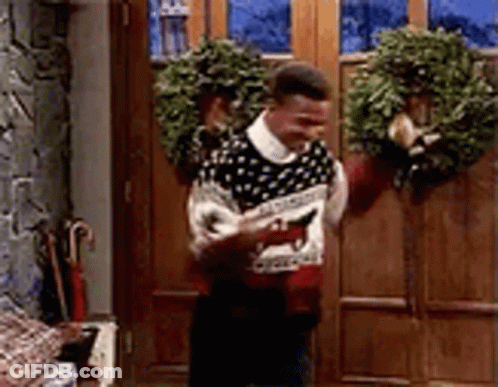 Man In Ugly Christmas Sweater Standing By Door With Wreath GIF