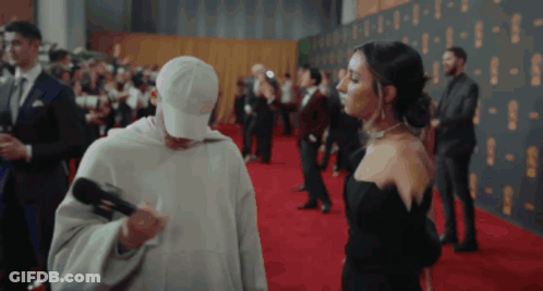 Man In White Hat Holding Microphone Next To Woman In Black Dress GIF