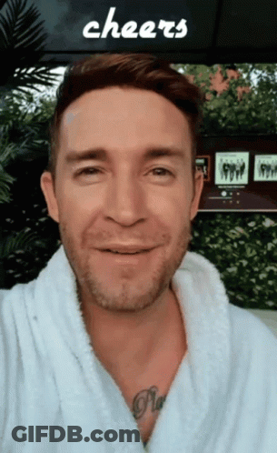 Man In White Robe Smiling In Front Of Cheers Sign GIF