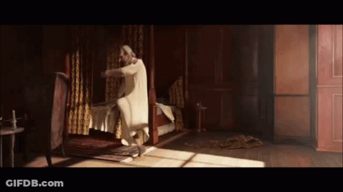 Man In White Robe Standing Next To Bed In Dark Room GIF