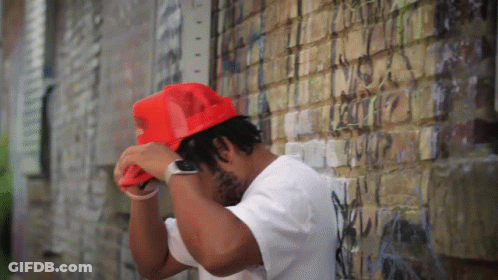 Man In White Shirt And Red Hat Leaning Against Brick Wall GIF