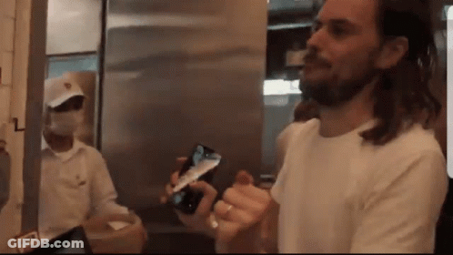 Man In White Shirt Holding Cell Phone In Front Of Masked Man GIF