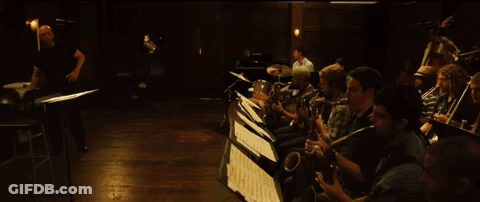 Man In White Shirt Kneeling Playing Drums In Dark Room GIF