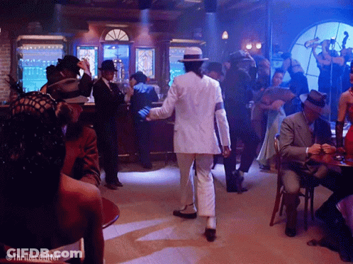 Man In White Suit Dancing In Room With People At Tables GIF