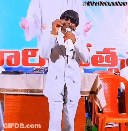 Man In White Suit Holding Microphone Before Mikelvelayudham Sign GIF