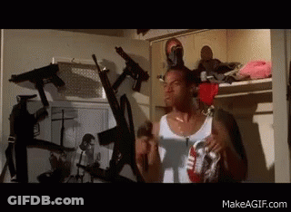 Man In White Tank Top Standing Surrounded By Guns GIF