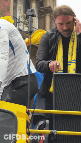 Man In Yellow Scarf With Letter E Standing In Front Of Speaker GIF