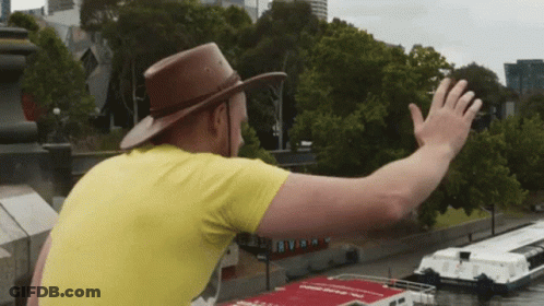 Man In Yellow Shirt And Brown Hat Waving At Boat GIF