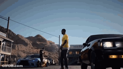 Man In Yellow Shirt Next To Black Car In Front Of Holiday Building GIF