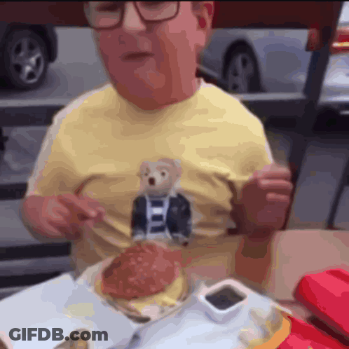Man In Yellow Shirt With Teddy Bear Eating Hamburger GIF