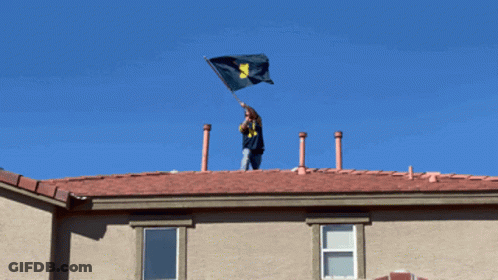 Man On Roof Holding Flag With Letter M GIF