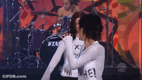 Man Playing Drums Behind Two Women, One Wearing Hate Shirt GIF