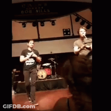 Man Playing Flute In Front Of Bob Mills Sign GIF