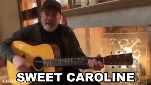 Man Playing Guitar In Front Of Fireplace Saying Sweet Caroline GIF