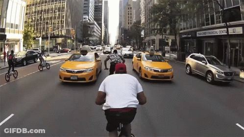 Man Riding Bike Down Street With Taxis And Bank Of America GIF