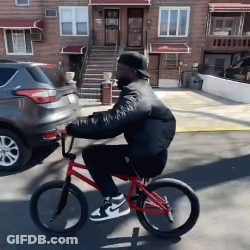Man Riding Red Bike Down Street Past Brick Building GIF