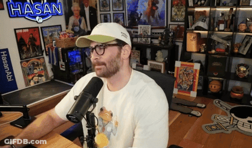 Man Sitting At Microphone With Sign Saying Hasan GIF