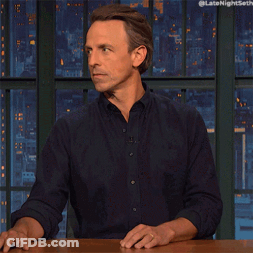 Man Sitting At Table With Hands Outstretched In Front Of Late Night Seth Screen GIF