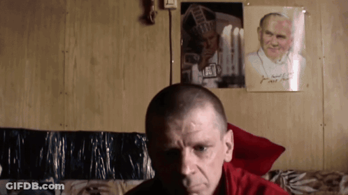 Man Sitting In Front Of Wall With Picture And John Paul Sign GIF