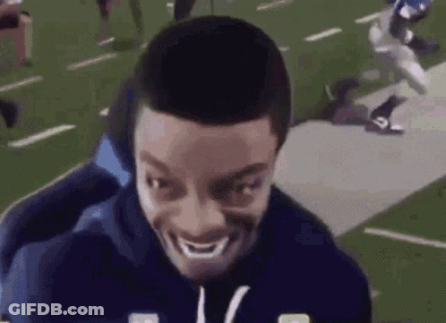 Man Smiling On Football Field With Player Running Behind GIF