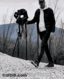 Man Walking Down Hill Holding Camera And Tripod GIF