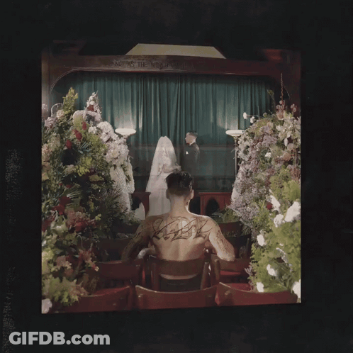 Man With Back Tattoo Beside Coffin And Flowers GIF