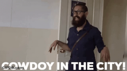 Man With Beard And Glasses Standing In Front Of Cowdoy Door GIF