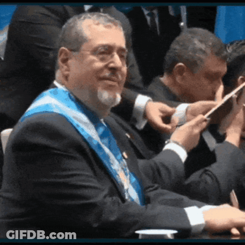Man With Glasses And Blue Sash Sitting At Table GIF