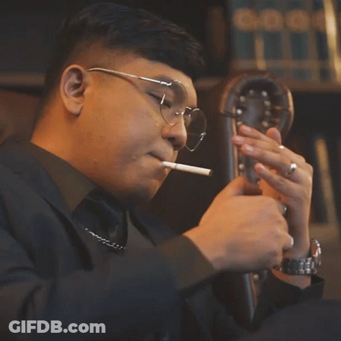Man With Glasses Smoking Cigarette In Front Of Bookshelf GIF