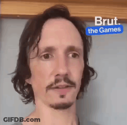 Man With Long Hair And Beard In Front Of Blue Sign Saying Brut The Games GIF