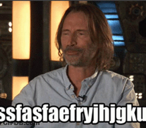 Man With Long Hair And Beard Sitting In Front Of SSFASFAEFRYJHJGKU Sign GIF
