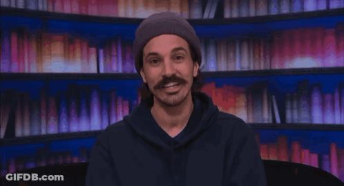 Man With Mustache In Beanie And Hoodie Smiling By Bookshelf GIF