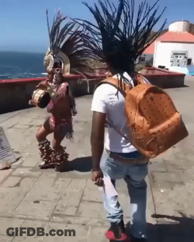 Man With Orange Mcm Backpack Next To Man In Native American Outfit GIF