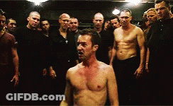 Men Standing Around Shirtless Man In Dark Room GIF