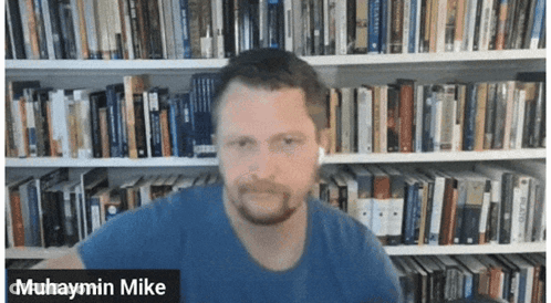 Muhaymin Mike Is Shown In Front Of A Library Full Of Books GIF