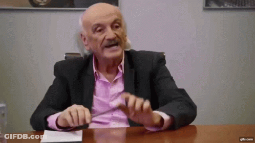 Older Man In Suit And Pink Shirt Talking At Table GIF