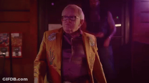 Older Man In Yellow Jacket And Glasses In Dark Room GIF