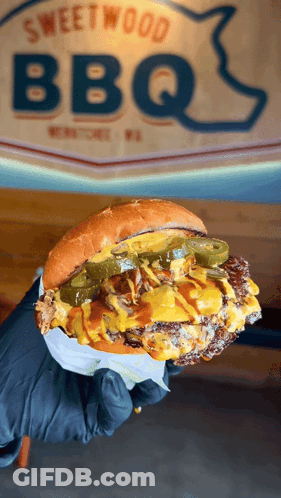 Person Holding Hamburger In Front Of Sweetwood BBQ Sign GIF