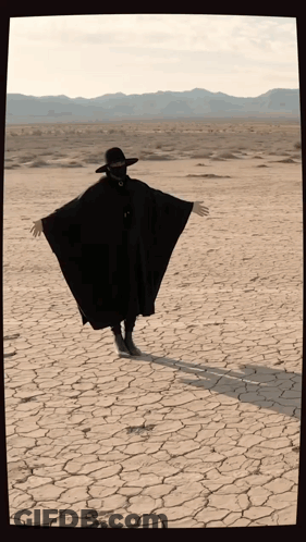 Person In Black Cape And Hat Arms Outstretched In Desert GIF